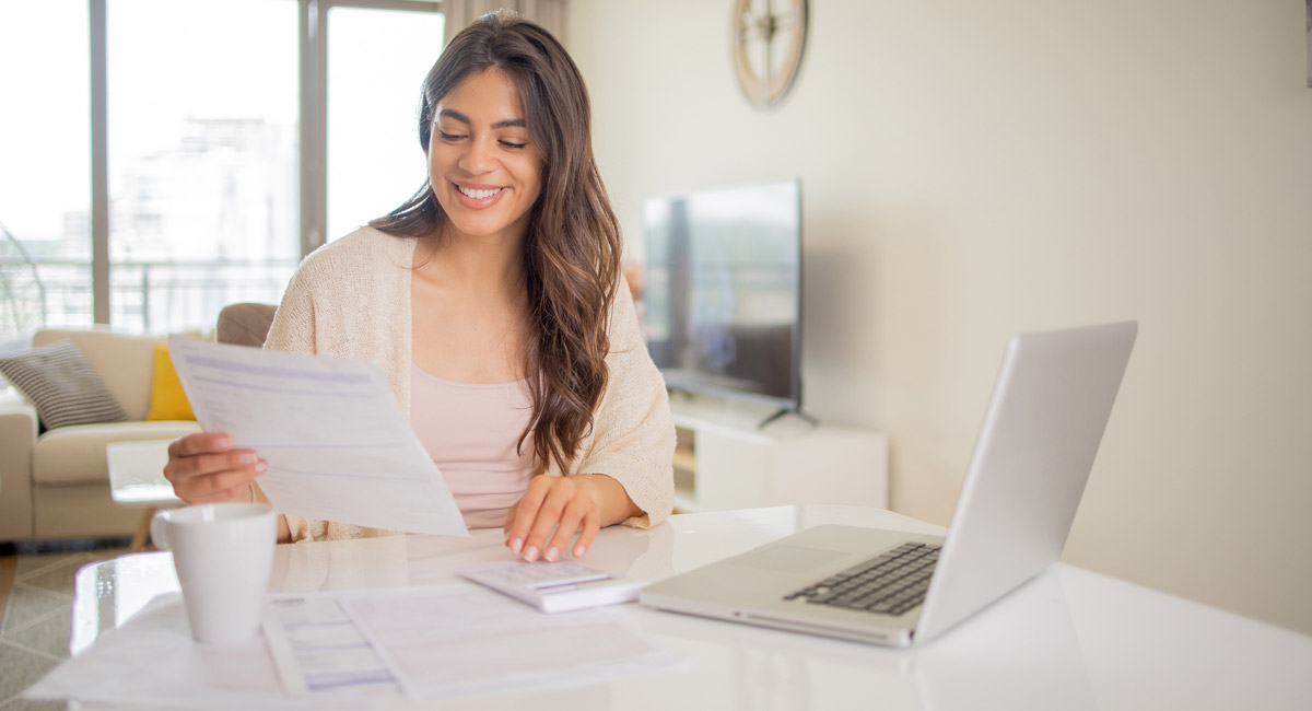 Woman smiling while reviewing papers at home with laptop